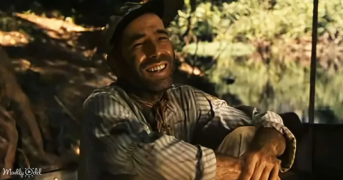A man in a striped shirt and hat is sitting outdoors, laughing with his head tilted back.