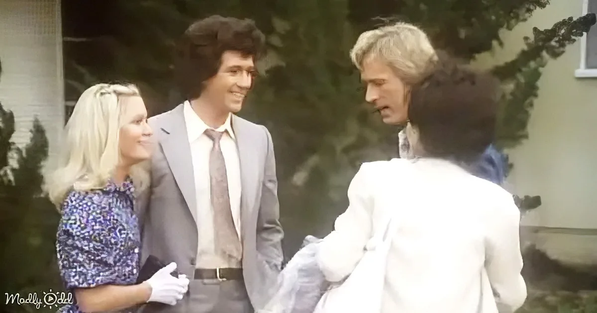 A man in a suit and tie is smiling while standing next to a woman in a floral dress, both looking at another man in a...