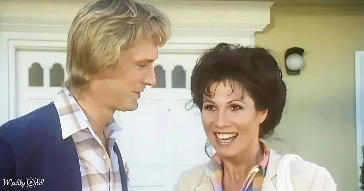 A smiling woman in a white jacket and a man in a blue suit are looking at each other in front of a white door.