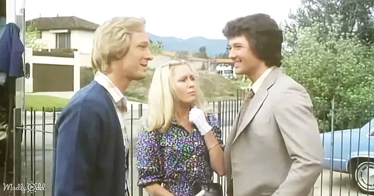 A woman in a colorful dress stands between two men, one in a blue suit and the other in a gray suit, in a suburban setting.
