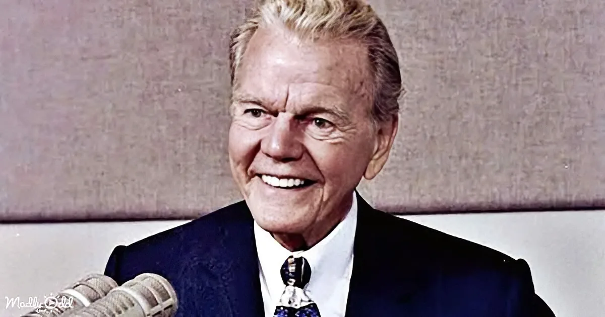 A smiling man in a suit and tie stands behind a microphone, likely hosting a radio or TV show.