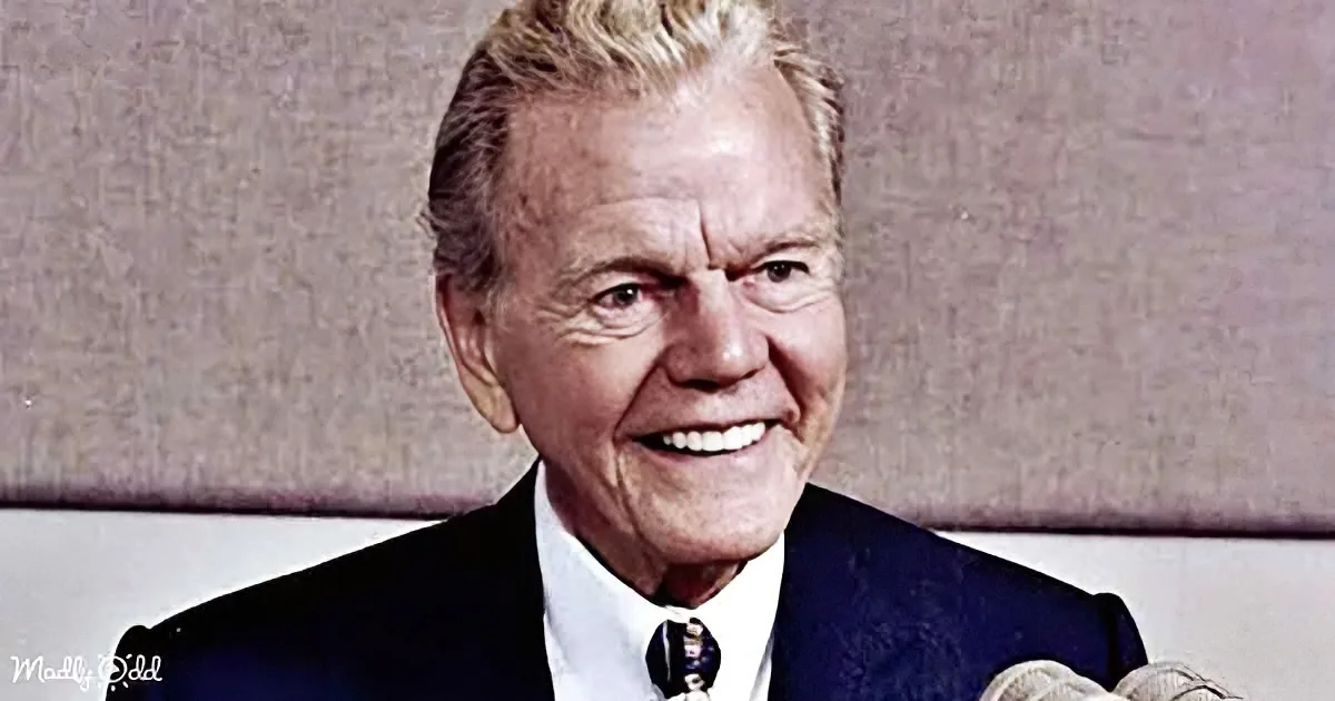 A smiling man with white hair, wearing a dark suit and tie, seated against a textured background.