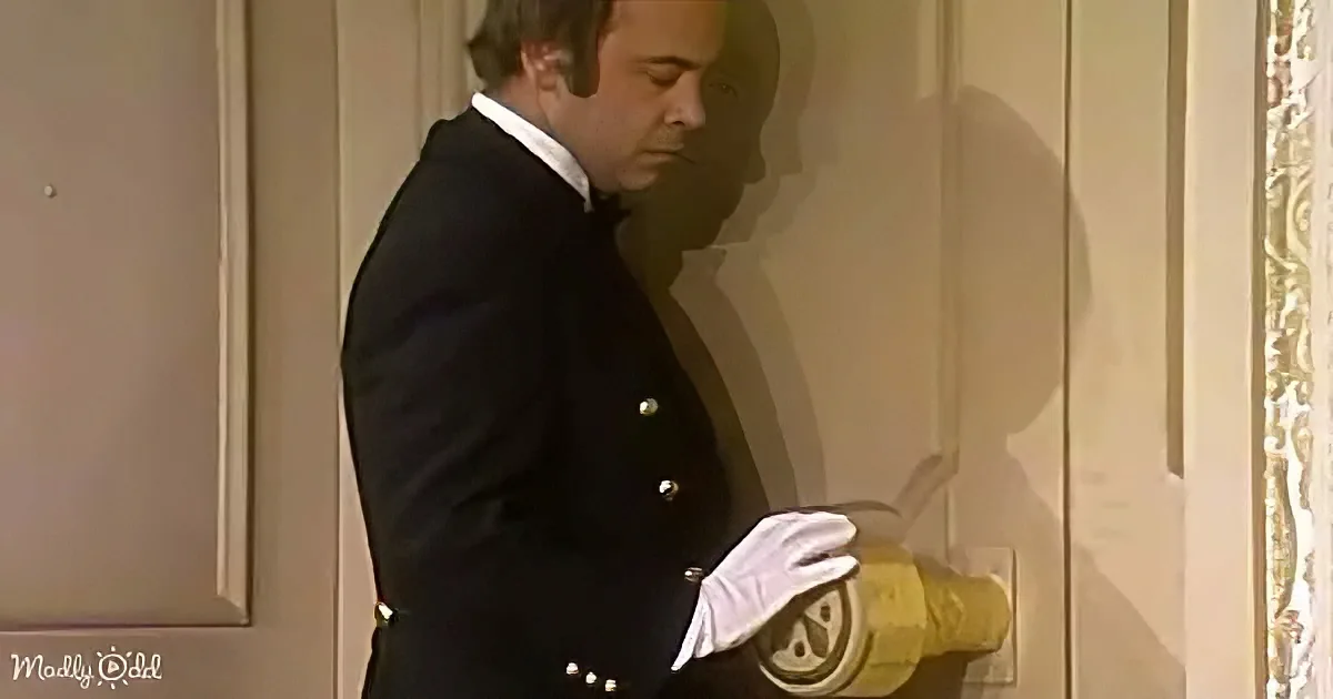 A man in a black suit and white gloves stands beside a door, holding a yellow object.