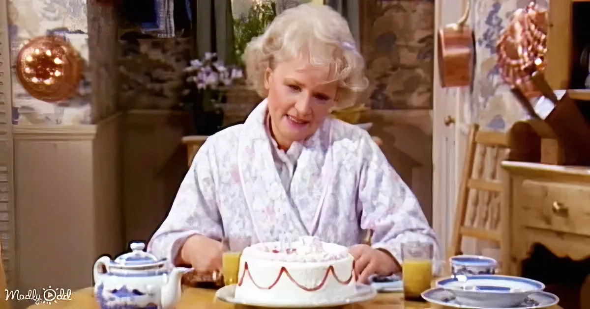 An elderly woman in a floral robe sits at a table with a cake, surrounded by tea cups and a teapot.