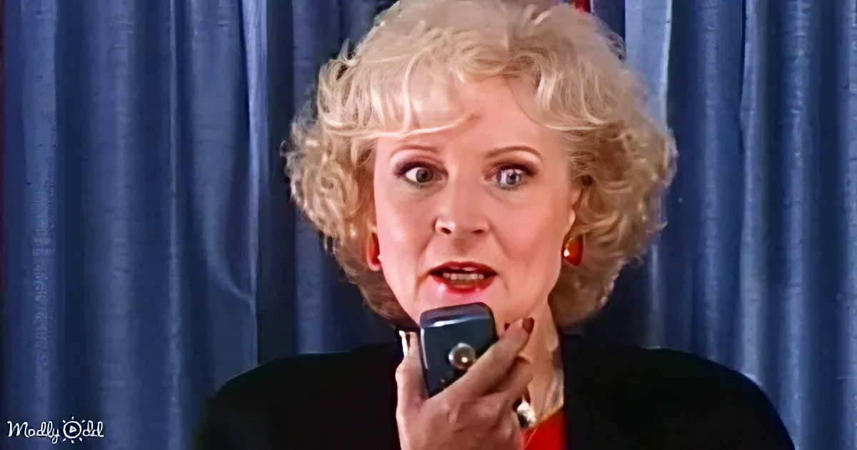 A woman with blonde, curly hair speaks into a blue microphone against a blue curtain backdrop.