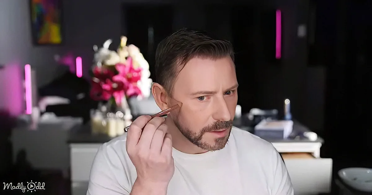 A man applies makeup with a brush, wearing a white shirt, in a room with pink lights and flowers.