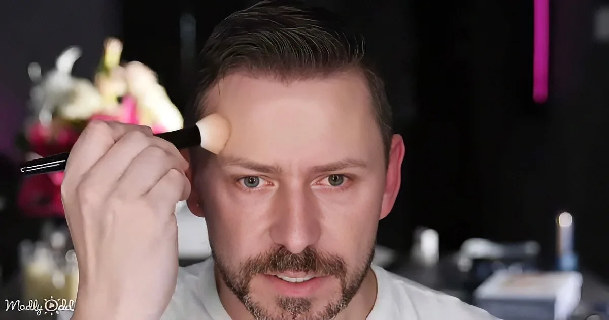 A man with a beard and mustache applies makeup with a brush, focusing on his face.