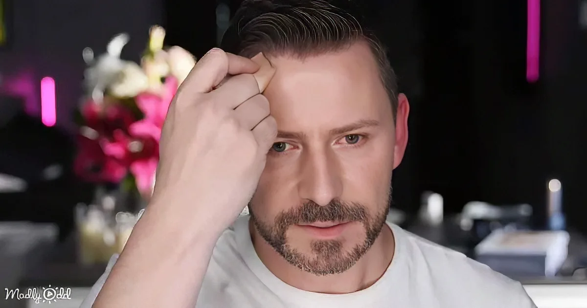 A man with a beard and short hair, wearing a white shirt, applies makeup with a brush near his forehead.