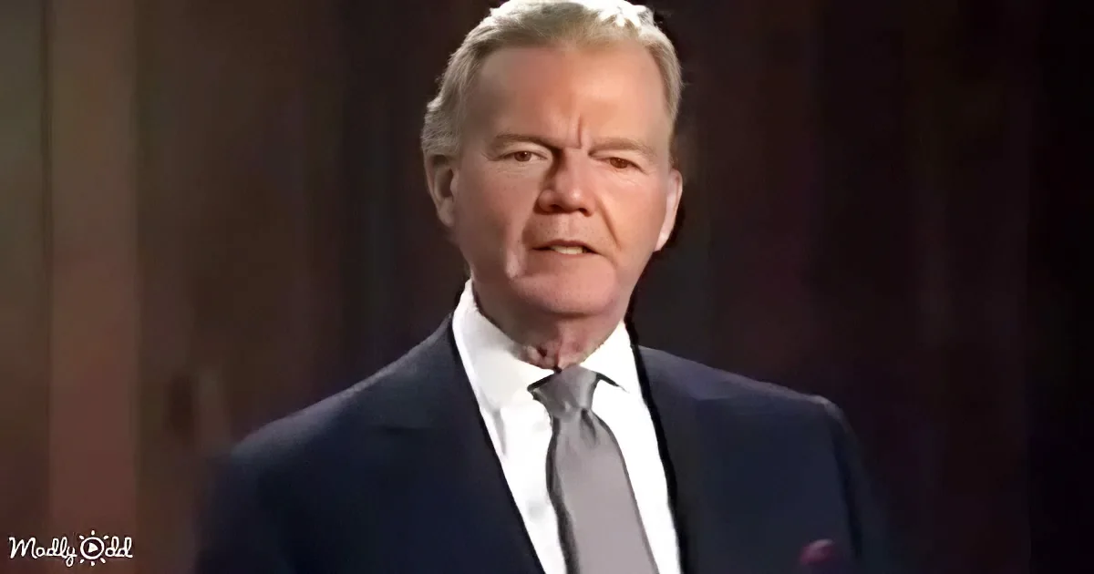 A man in a dark suit and white shirt with a gray tie stands against a dark background.