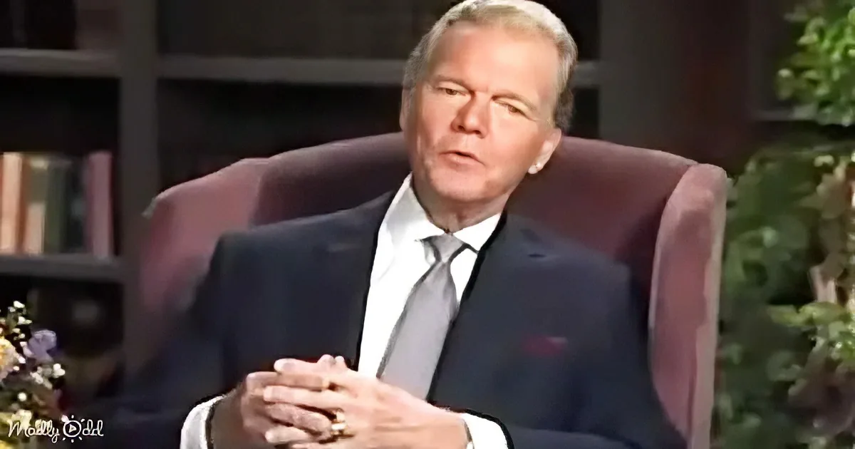 A man in a suit sits in a chair, hands clasped, with a bookshelf and greenery in the background.