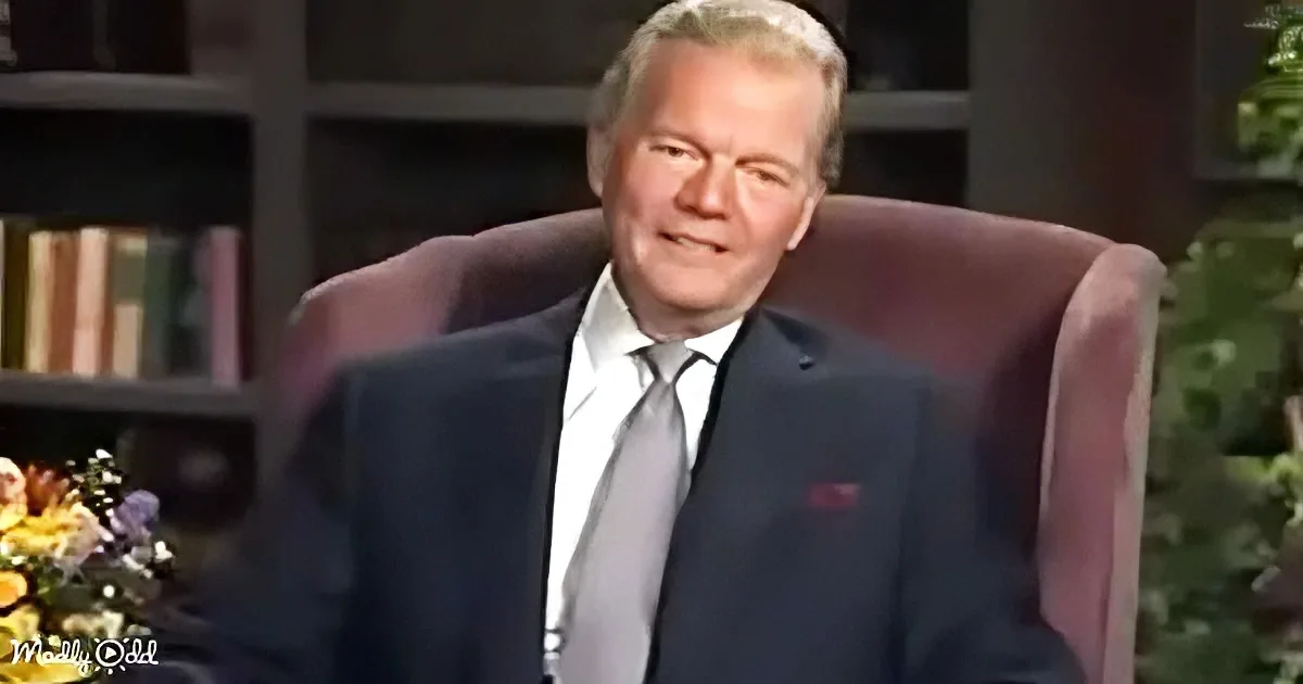 A man in a suit sits in a chair, smiling, with a bookshelf and flowers in the background.
