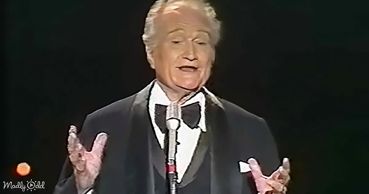 A man in a tuxedo and bow tie is speaking on stage with his hands raised.