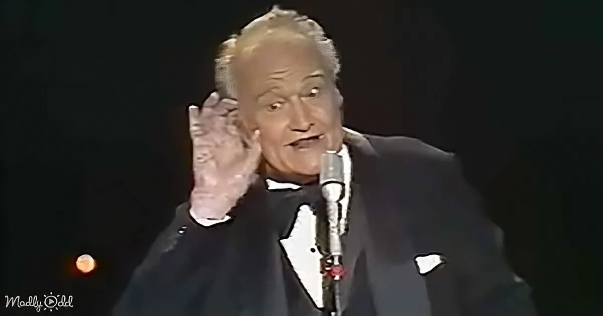 A man in a tuxedo gestures with his right hand while speaking into a microphone.