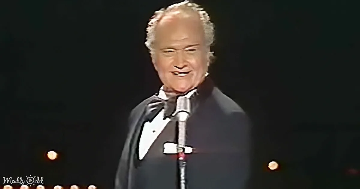 A man in a tuxedo smiles on stage, holding a microphone, with a spotlight shining on him.