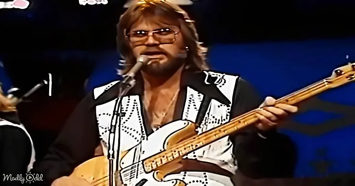 A man with glasses and a beard plays a guitar on stage, wearing a white and black patterned shirt.