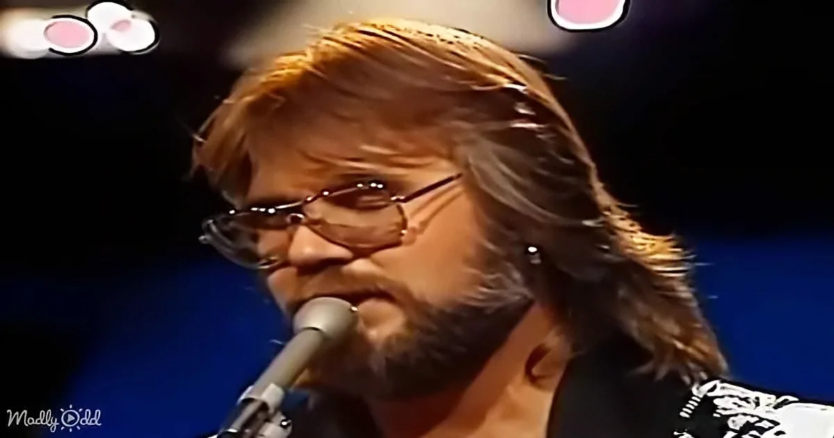 A person with long hair and glasses sings into a microphone on stage.