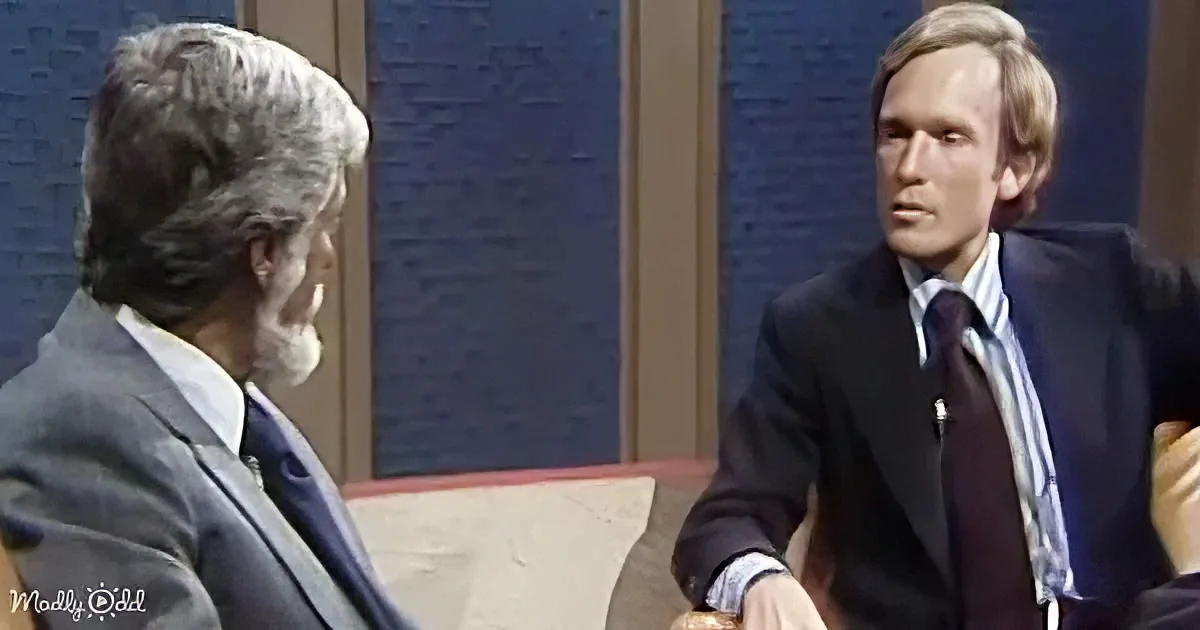 A man in a suit sits on a couch, engaged in conversation with another man in a suit, both appearing serious.