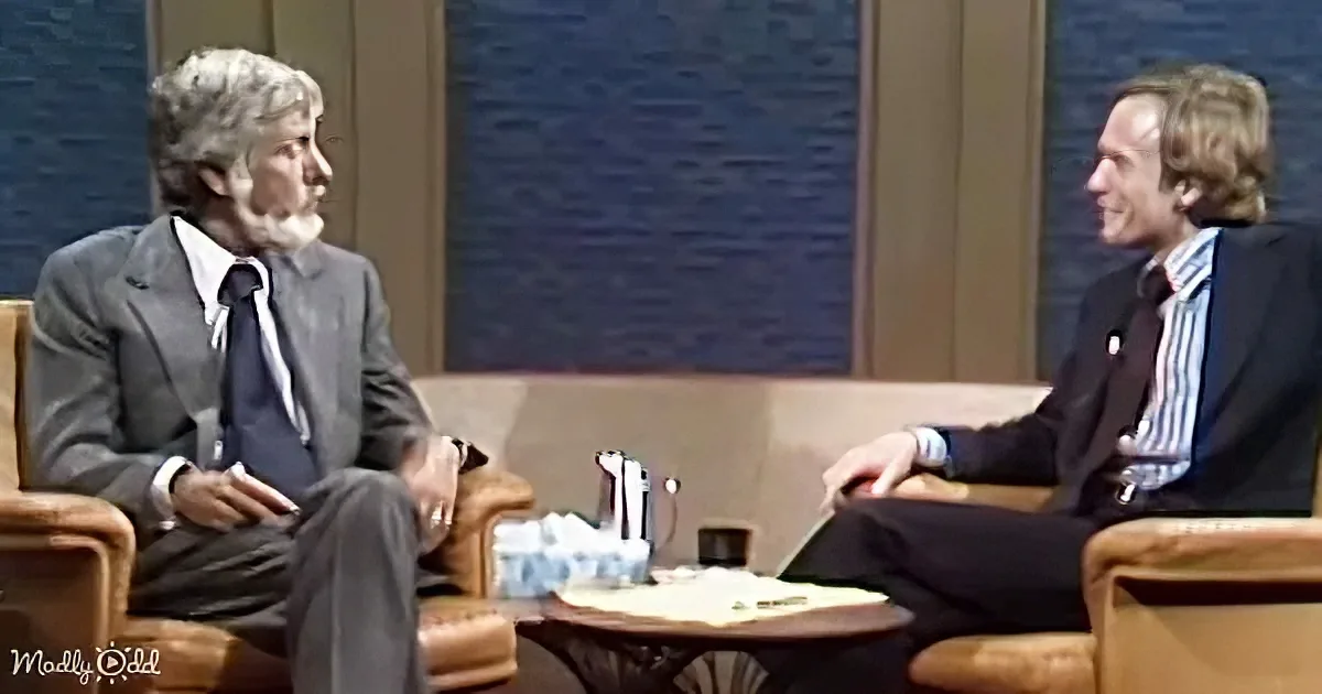 Two men in suits sit on couches, engaged in conversation, with a table and tissues between them.