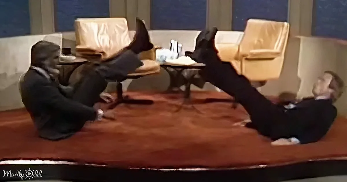 Two men in suits are sitting on a stage, one with legs up, both appearing relaxed.