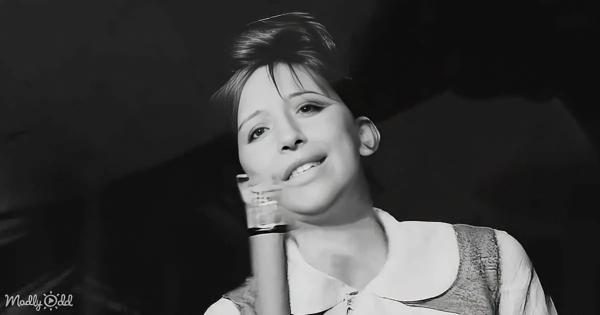 A person holding a microphone, smiling, wearing a sleeveless top, in a dimly lit setting.