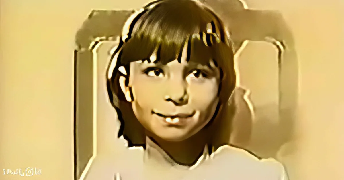 A young boy with short hair and bangs, wearing a white shirt, is smiling and looking directly at the camera.
