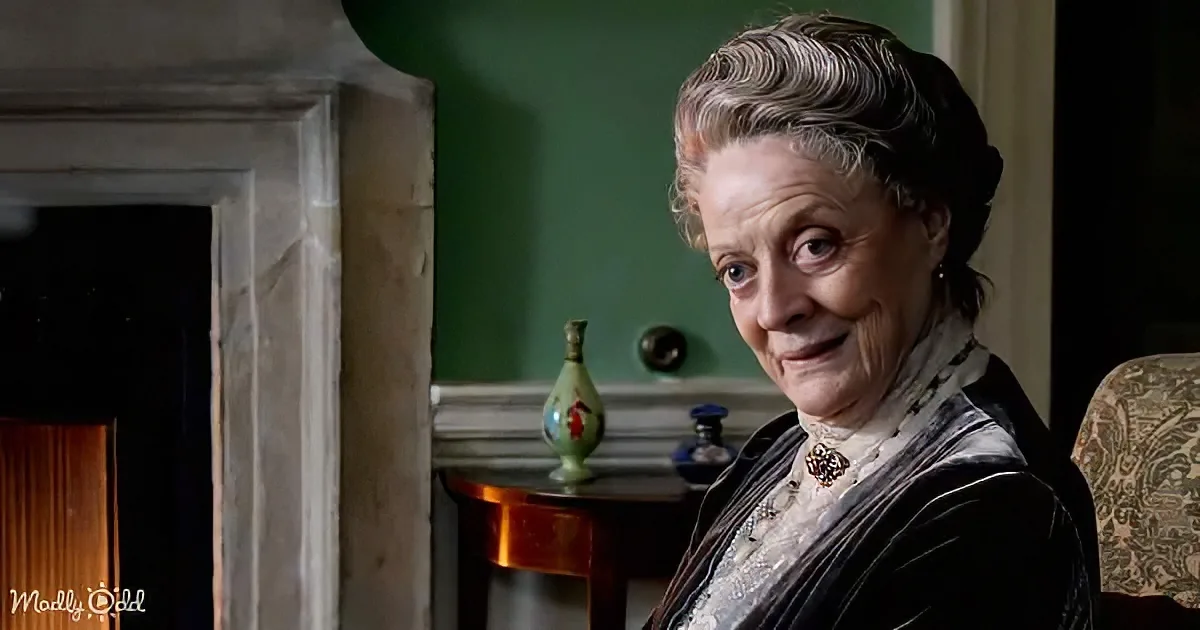 An elderly woman with gray hair, wearing a black dress and pearl necklace, sits in a room with green walls and a fireplace.