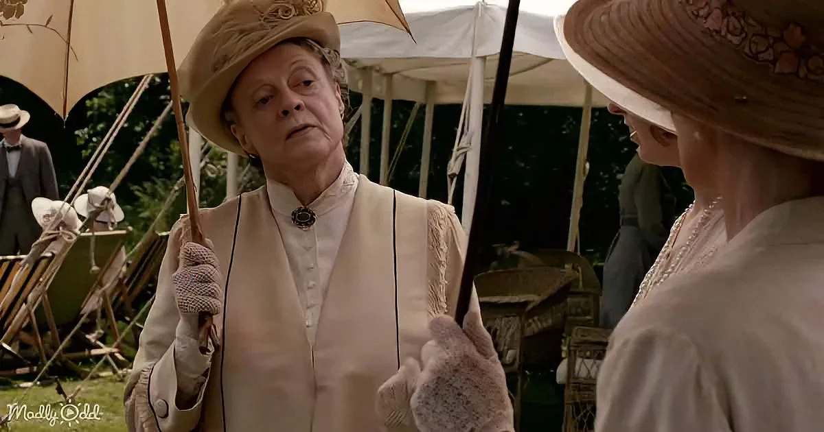 An older woman in a white Victorian-style outfit holds a parasol, standing outdoors with another woman in period attire.