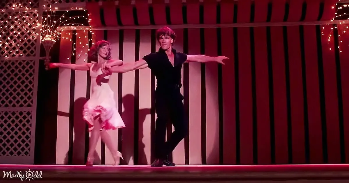 A man and woman dance gracefully on stage, arms outstretched, under warm red lighting.