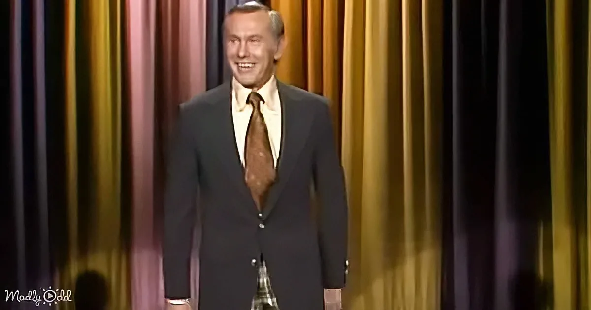 A man in a dark suit and brown tie smiles on stage with a colorful curtain backdrop.