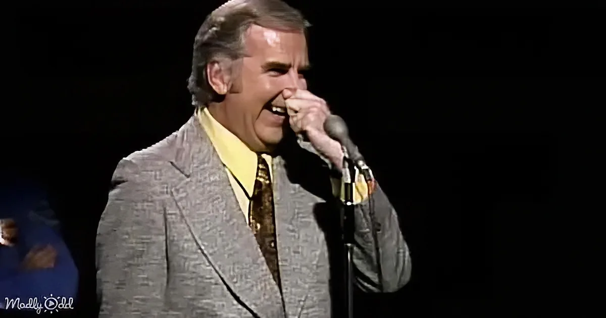 A man in a suit sings into a microphone on stage, wearing a yellow shirt and patterned tie.