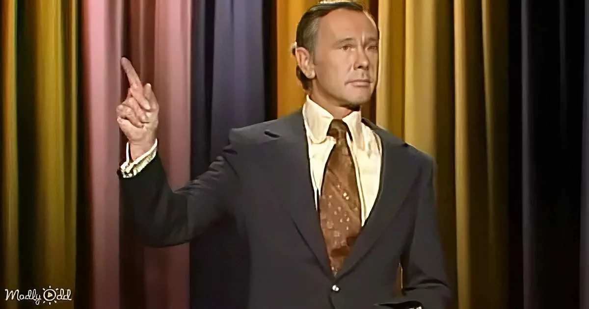 A man in a suit and tie points upwards, standing against a colorful curtain backdrop.
