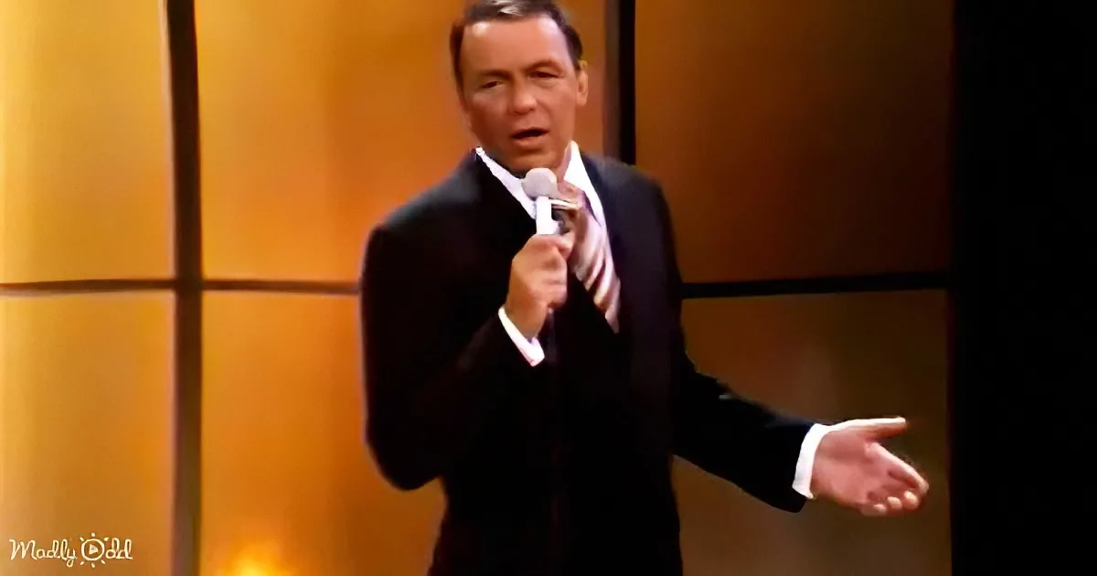 A man in a black suit holds a microphone, gesturing with his right hand, against a warm-toned backdrop.