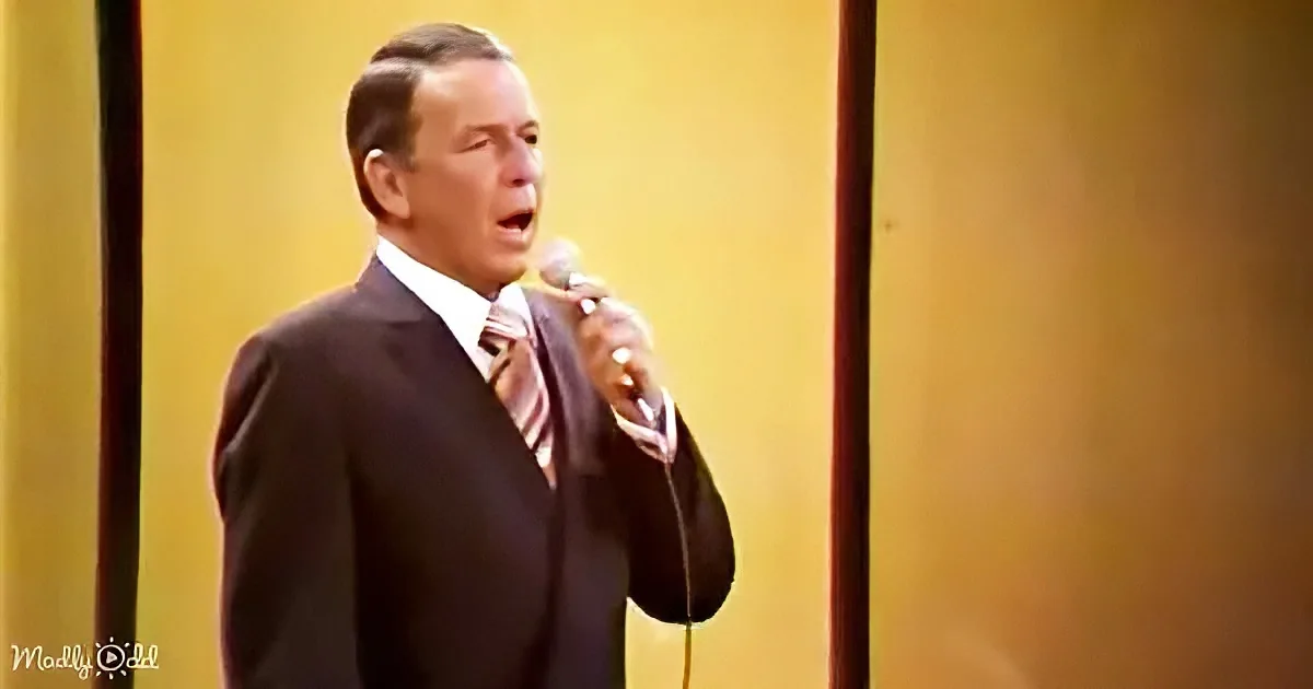 A man in a suit sings into a microphone against a yellow background.