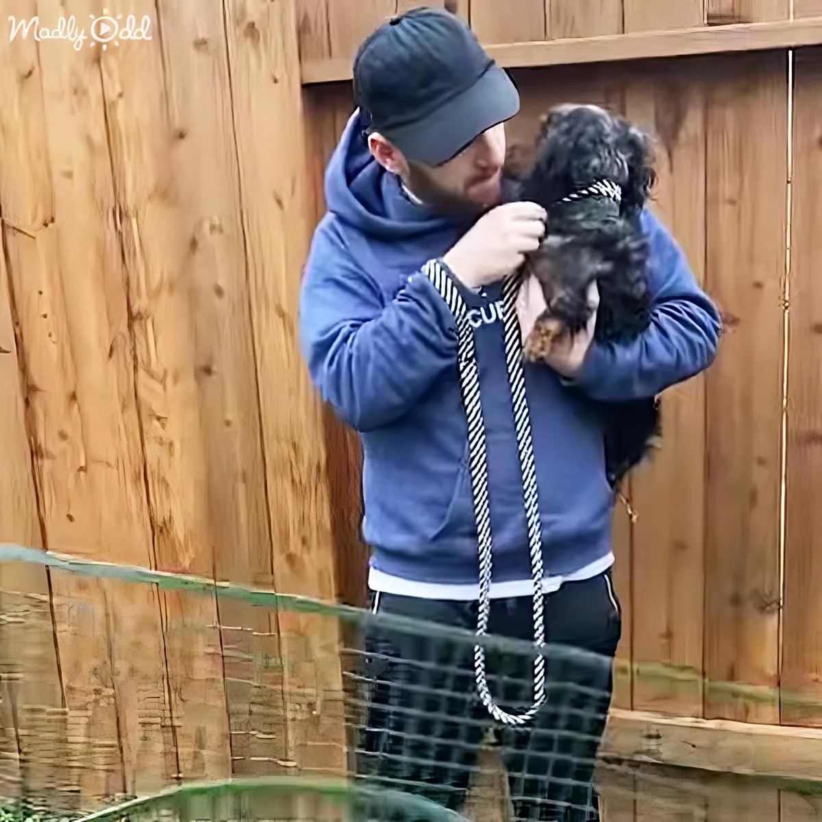 A man in a blue hoodie and beanie holds a small dog with a rope leash, standing in front of a wooden fence.