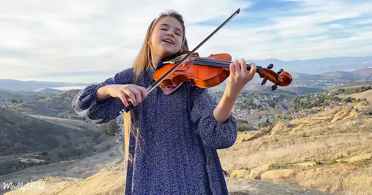 A girl in a blue dress plays the violin on a hilltop with a scenic view of mountains and a town below.