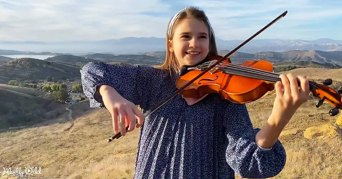 A girl in a blue dress plays a violin on a hillside with a scenic view of mountains and a lake in the background.