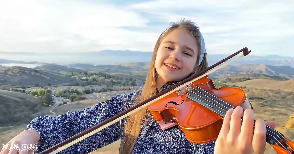 A girl smiles while playing the violin against a scenic backdrop of hills and a distant cityscape.