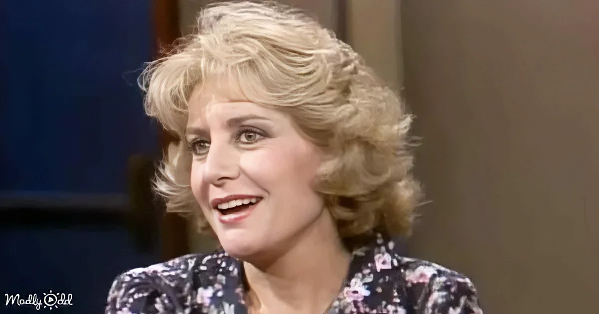 A woman with blonde, shoulder-length hair smiles while wearing a floral-patterned blouse.