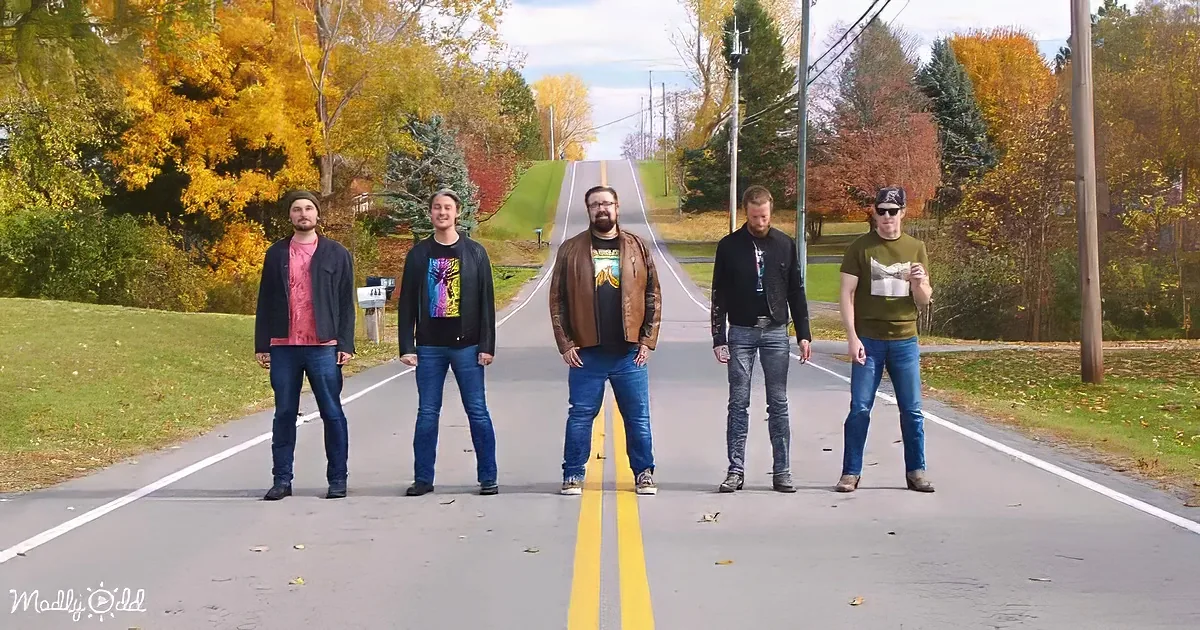 Five men stand on a road with autumn foliage and a clear sky in the background.