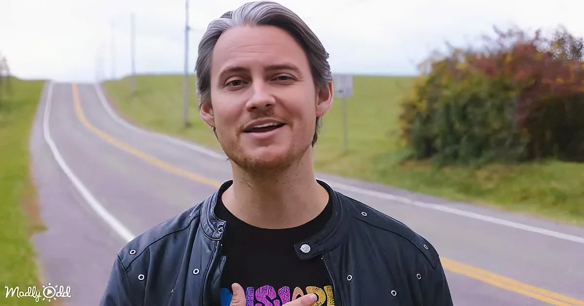 A man in a black leather jacket and t-shirt stands on a road with a scenic background.
