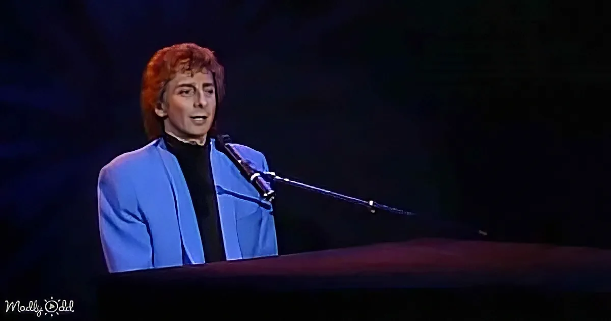 A person in a blue suit sings into a microphone on stage, with a dark background.