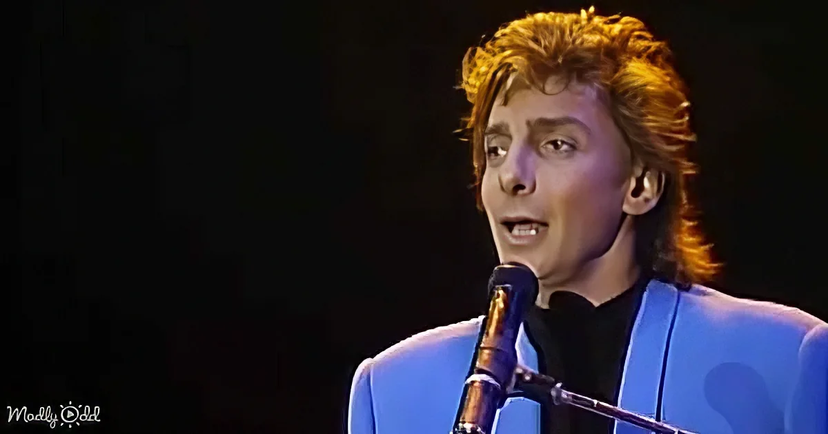 A person with curly hair in a blue suit sings into a microphone against a dark background.