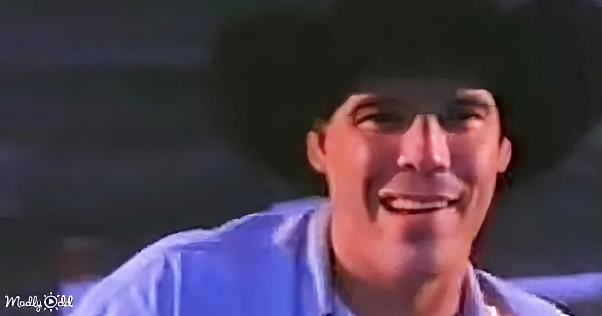 A man in a blue shirt and cowboy hat is smiling, with a blurred background.