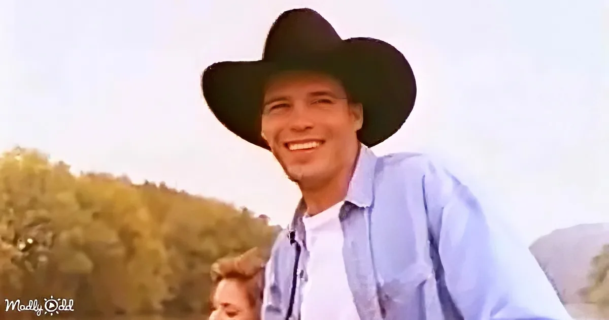 A man in a cowboy hat and blue shirt smiles, with a blurred background of trees and sky.