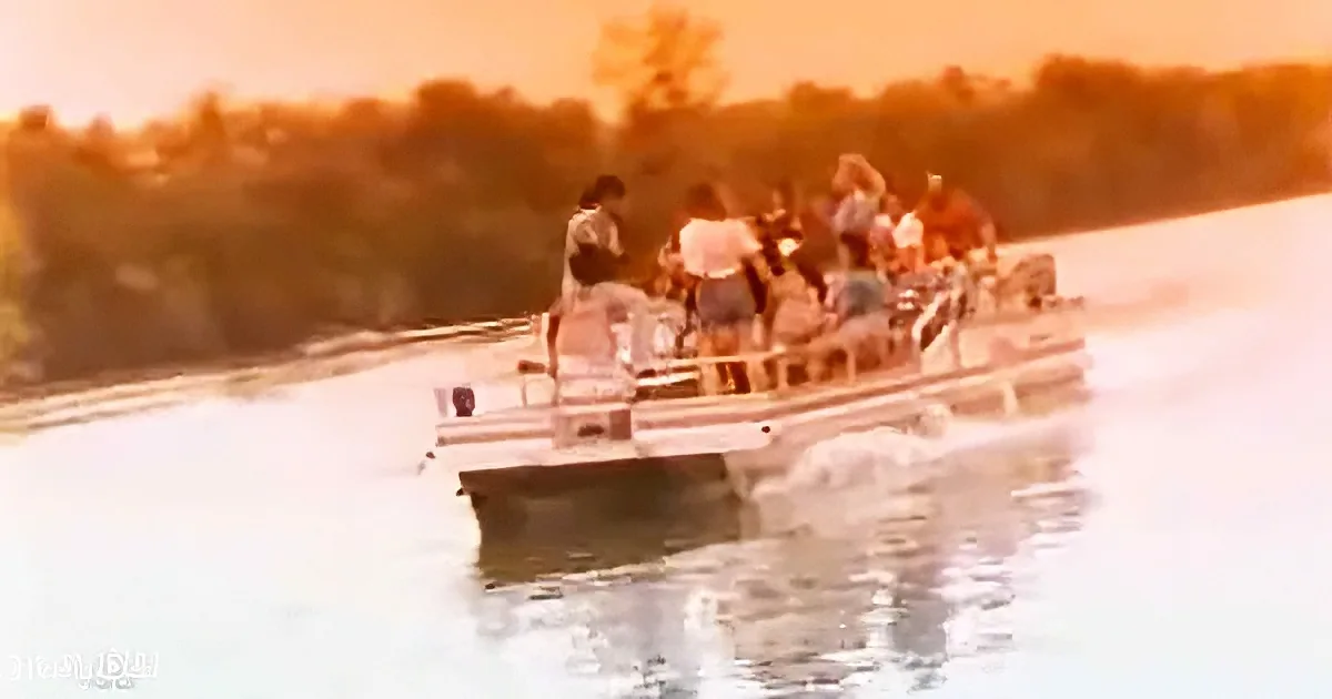A pontoon boat with people on it is moving across a body of water with trees in the background.