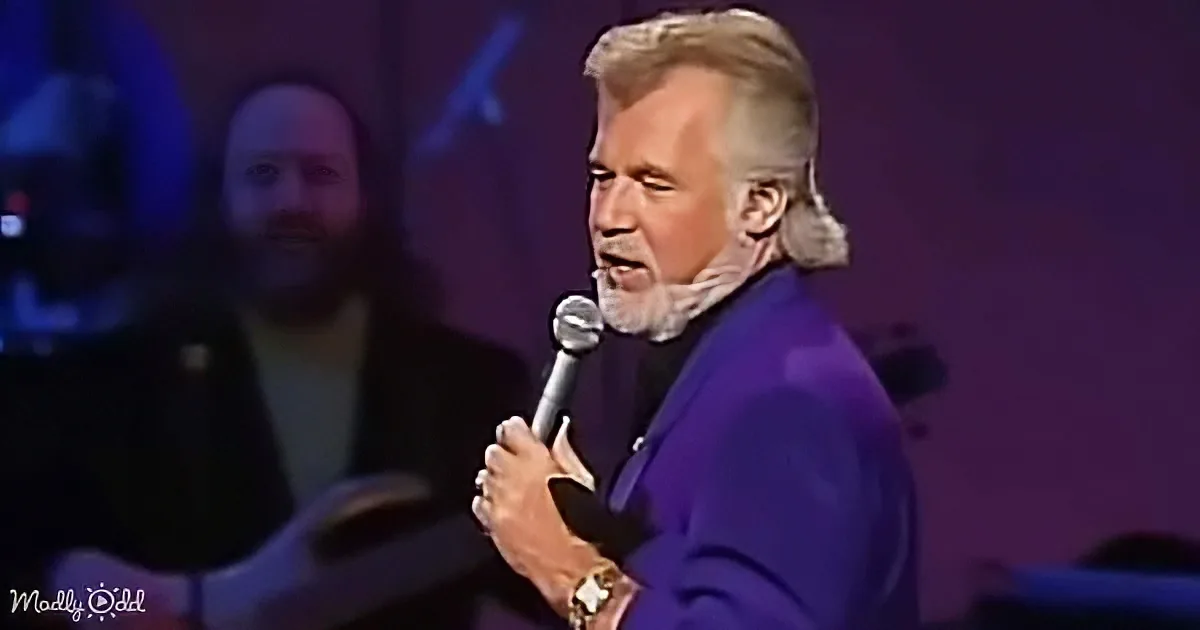 A man in a purple suit sings into a microphone, with a blurred background and a "Madly Odd" watermark.
