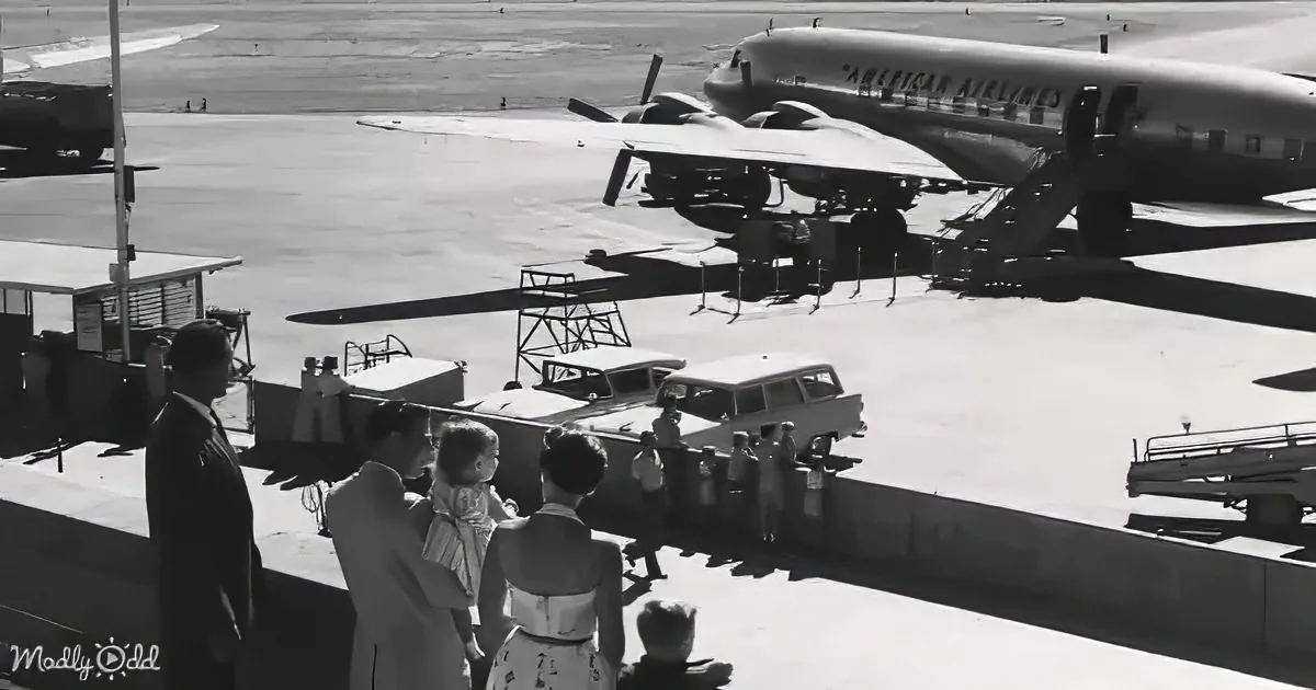 A vintage scene at an airport with people watching a parked American Airlines plane and a car being loaded onto a ramp.
