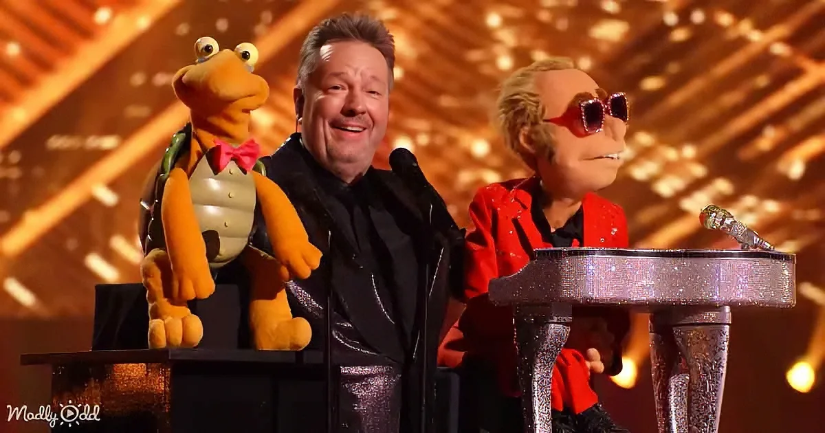 A man stands between two puppets, one resembling a turtle and the other a person in red, on a stage with a glittery...