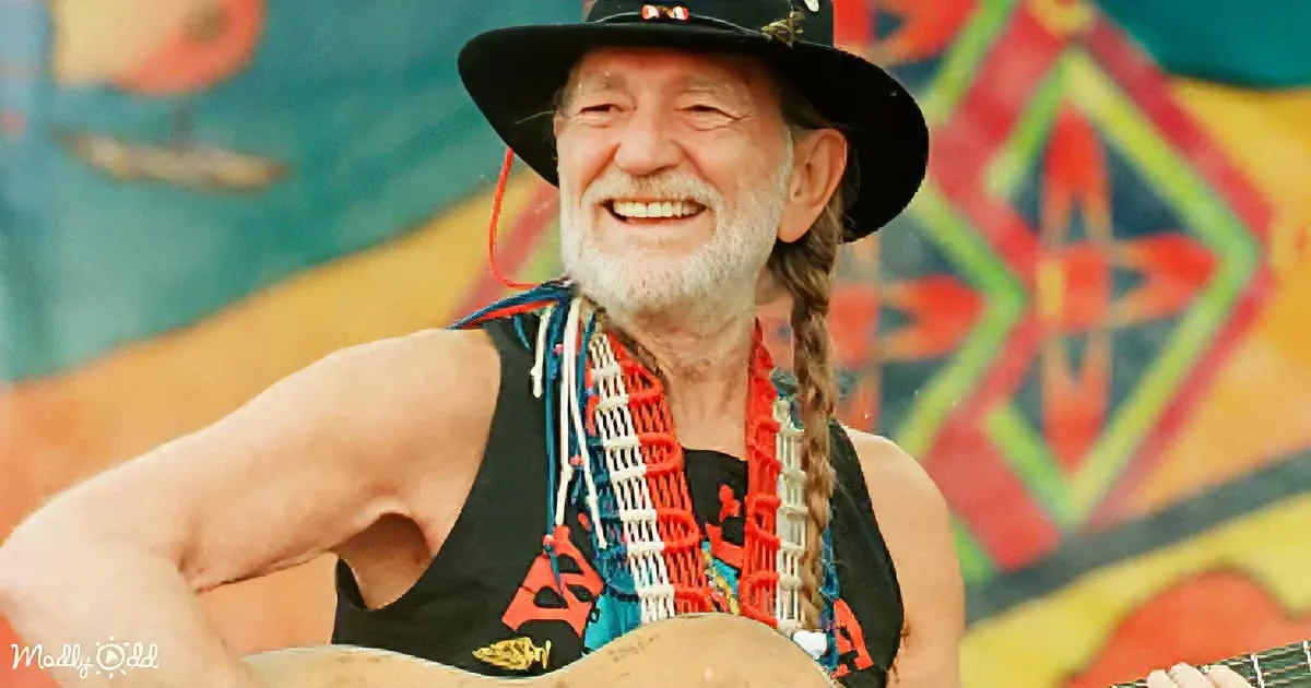 A smiling man with braided hair, wearing a black hat and a colorful necklace, holds a guitar in front of a vibrant mural.
