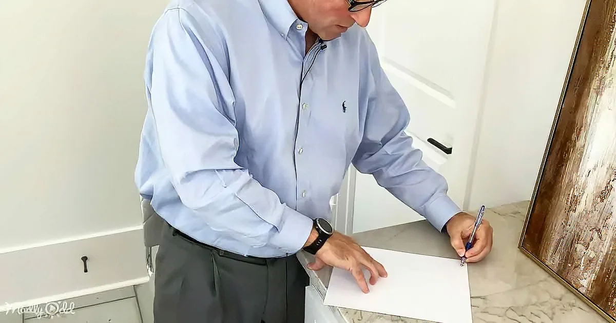 A man in a blue shirt and glasses writes on a white paper with a pen.